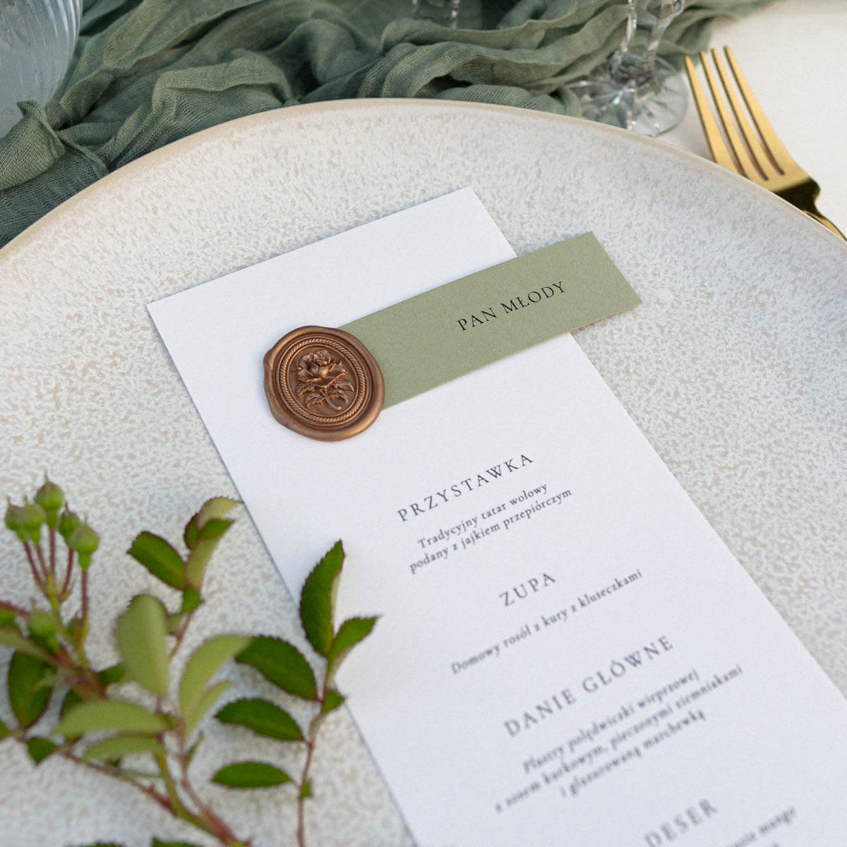 Menu ślubne z winietką na eleganckim papierze, ozdobione złotą pieczęcią lakową i subtelnymi detalami dekoracyjnymi, ułożone na tle materiału i kwiatów. Papeteria ślubna w stylu eleganckim, idealna dekoracja stołu weselnego i praktyczny dodatek na wesele.