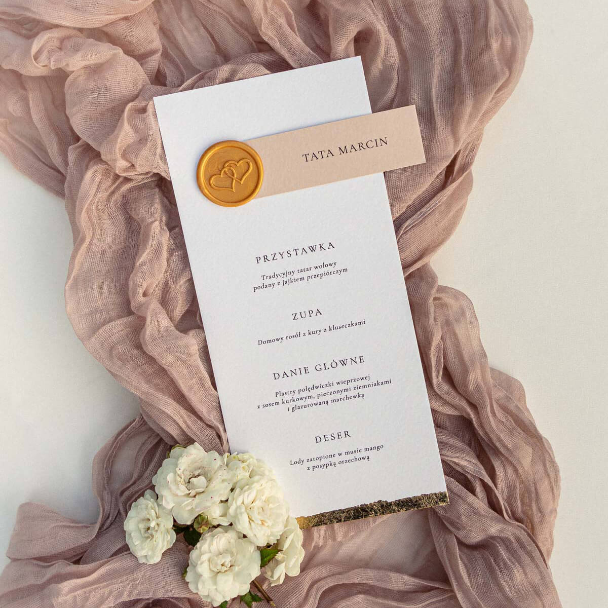 Menu ślubne z winietką na eleganckim papierze, ozdobione złotą pieczęcią lakową i subtelnymi detalami dekoracyjnymi, ułożone na tle materiału i kwiatów. Papeteria ślubna w stylu eleganckim, idealna dekoracja stołu weselnego i praktyczny dodatek na wesele.
