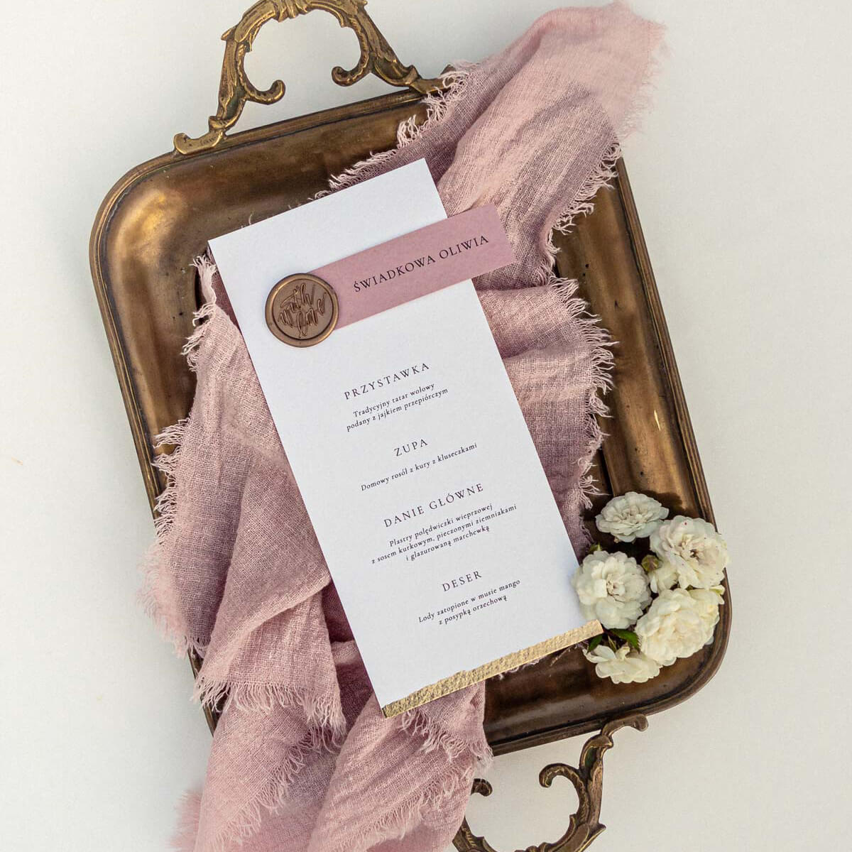Menu ślubne z winietką na eleganckim papierze, ozdobione złotą pieczęcią lakową i subtelnymi detalami dekoracyjnymi, ułożone na tle materiału i kwiatów. Papeteria ślubna w stylu eleganckim, idealna dekoracja stołu weselnego i praktyczny dodatek na wesele.