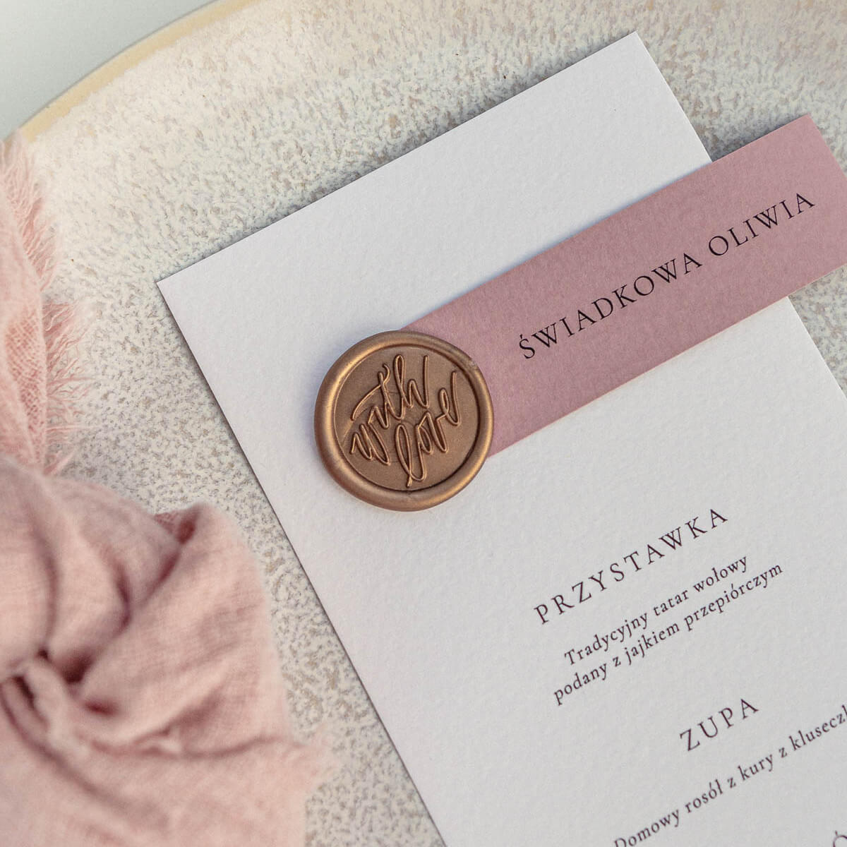 Menu ślubne z winietką na eleganckim papierze, ozdobione złotą pieczęcią lakową i subtelnymi detalami dekoracyjnymi, ułożone na tle materiału i kwiatów. Papeteria ślubna w stylu eleganckim, idealna dekoracja stołu weselnego i praktyczny dodatek na wesele.