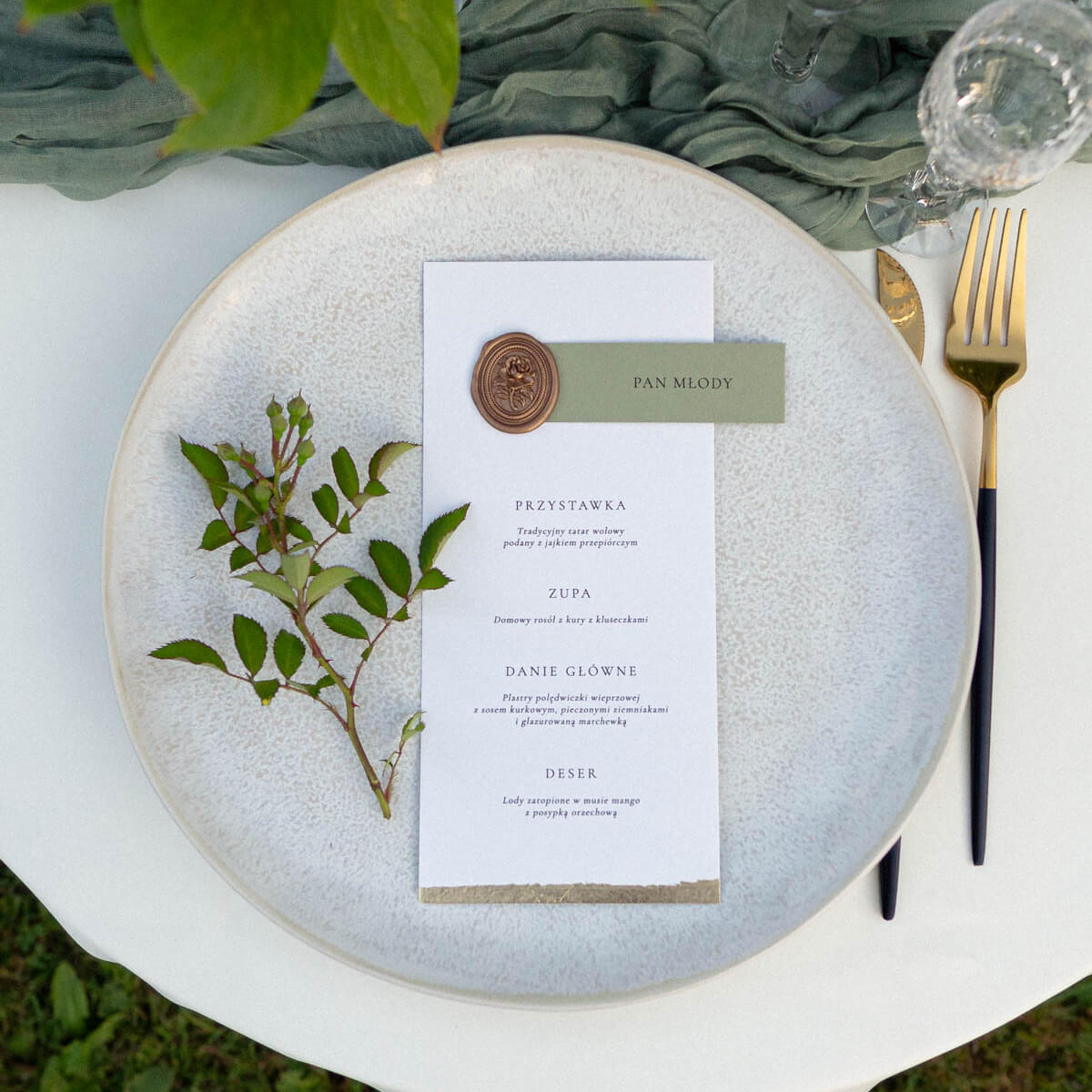 Menu ślubne z winietką na eleganckim papierze, ozdobione złotą pieczęcią lakową i subtelnymi detalami dekoracyjnymi, ułożone na tle materiału i kwiatów. Papeteria ślubna w stylu eleganckim, idealna dekoracja stołu weselnego i praktyczny dodatek na wesele.