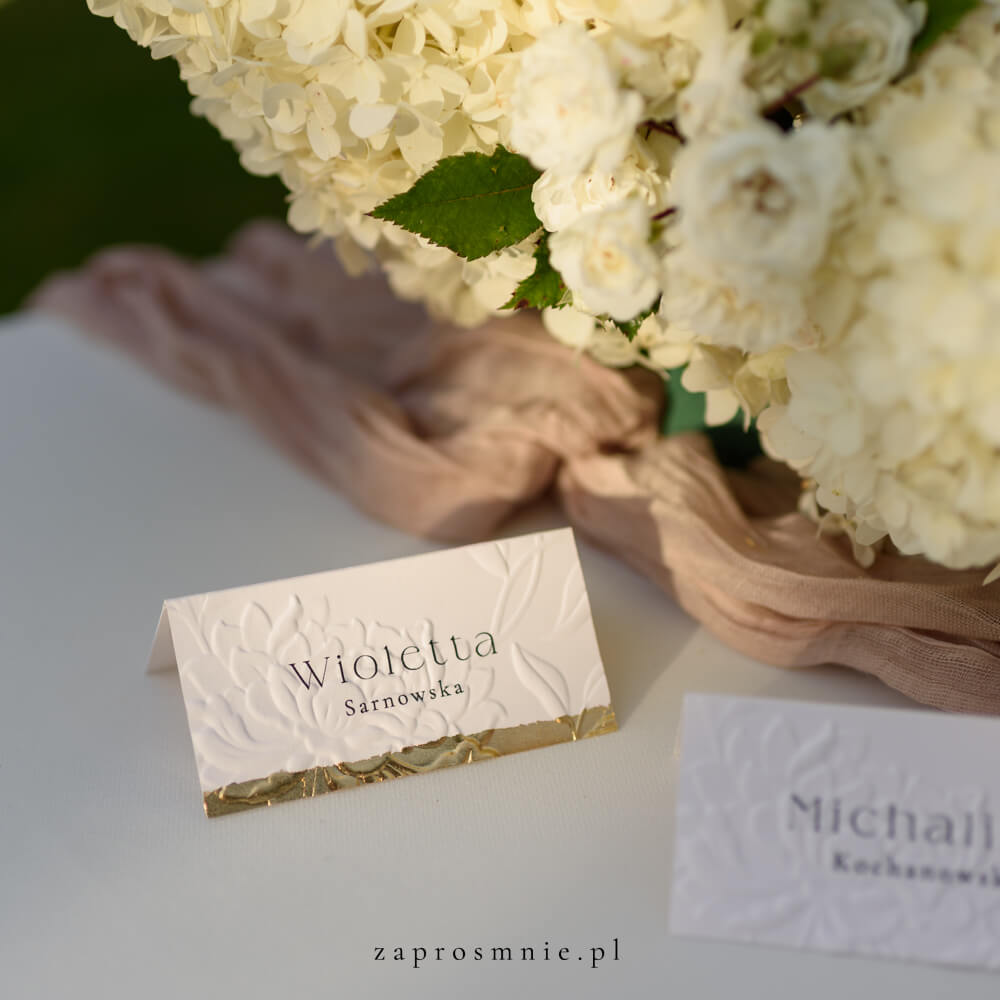 winietka ślubna z eleganckim tłoczeniem kwiatowym i złotym wykończeniem na dole, z imieniem i nazwiskiem gościa, ustawiona na stole obok kwiatowej dekoracji i tkaniny w pastelowym odcieniu, elegancka papeteria ślubna, która ułatwia organizację miejsc i dodaje klasy aranżacji weselnej, winietka z tłoczeniem