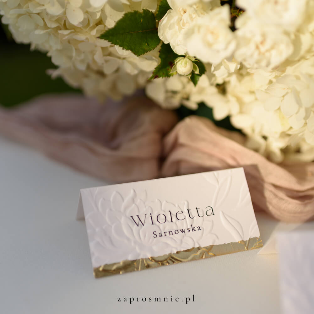 winietka ślubna z eleganckim tłoczeniem kwiatowym i złotym wykończeniem na dole, z imieniem i nazwiskiem gościa, ustawiona na stole obok kwiatowej dekoracji i tkaniny w pastelowym odcieniu, elegancka papeteria ślubna, która ułatwia organizację miejsc i dodaje klasy aranżacji weselnej, winietka z tłoczeniem