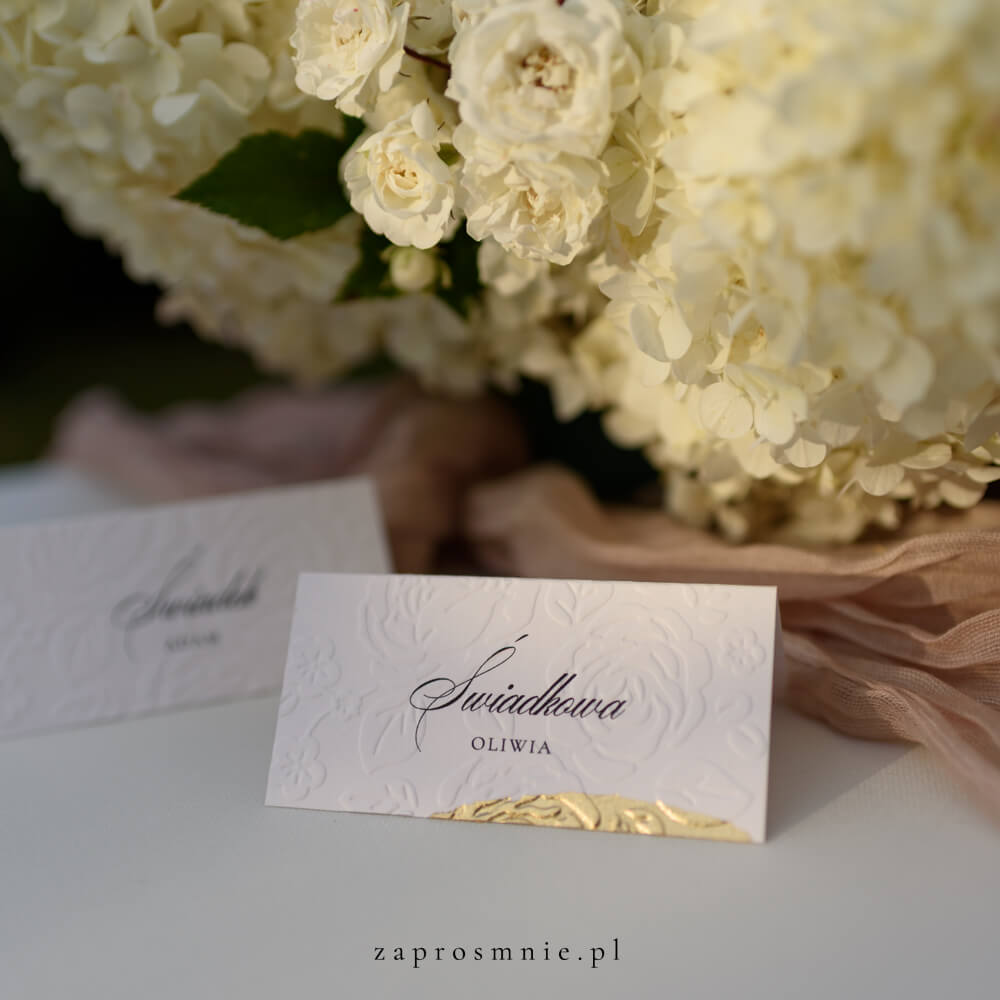 winietka ślubna z eleganckim tłoczeniem kwiatowym i złotym wykończeniem na dole, z imieniem i nazwiskiem gościa, ustawiona na stole obok kwiatowej dekoracji i tkaniny w pastelowym odcieniu, elegancka papeteria ślubna, która ułatwia organizację miejsc i dodaje klasy aranżacji weselnej, winietka z tłoczeniem