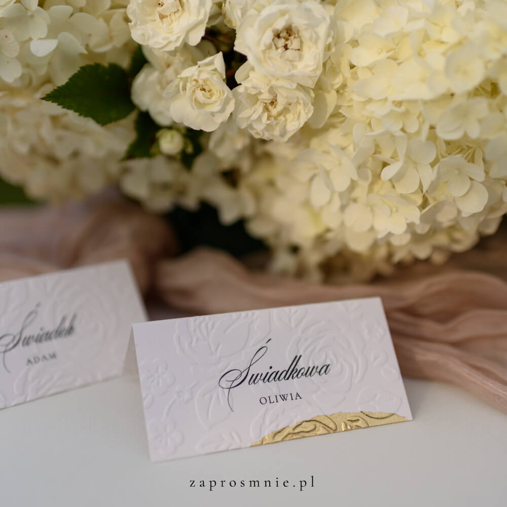 winietka ślubna z eleganckim tłoczeniem kwiatowym i złotym wykończeniem na dole, z imieniem i nazwiskiem gościa, ustawiona na stole obok kwiatowej dekoracji i tkaniny w pastelowym odcieniu, elegancka papeteria ślubna, która ułatwia organizację miejsc i dodaje klasy aranżacji weselnej, winietka z tłoczeniem