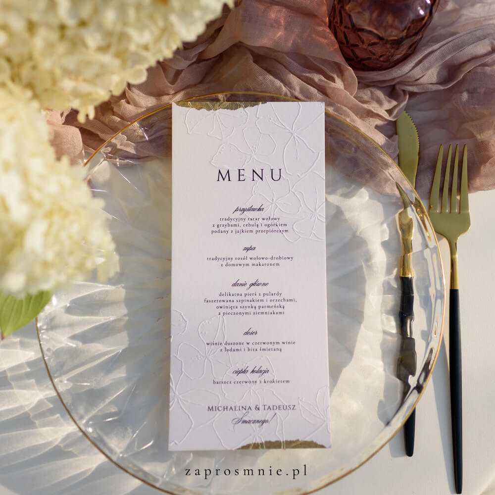 Ekskluzywne menu weselne w bieli z eleganckim tłoczeniem roślinnym i złotym wykończeniem na brzegach, położone na przezroczystym talerzu ze złotymi sztućcami, luksusowa papeteria ślubna w stylu glamour, która dodaje klasy aranżacji stołu i podkreśla wyjątkowy charakter przyjęcia weselnego, menu z tłoczeniem kwiatowym