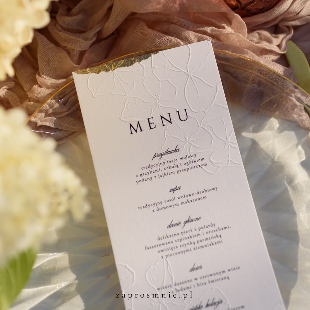 Ekskluzywne menu weselne w bieli z eleganckim tłoczeniem roślinnym i złotym wykończeniem na brzegach, położone na przezroczystym talerzu ze złotymi sztućcami, luksusowa papeteria ślubna w stylu glamour, która dodaje klasy aranżacji stołu i podkreśla wyjątkowy charakter przyjęcia weselnego, menu z tłoczeniem kwiatowym