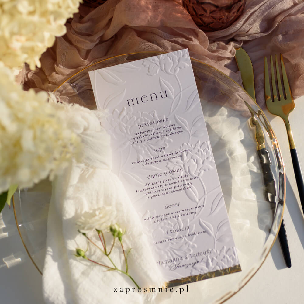 menu weselne z eleganckim tłoczeniem kwiatowym, położone na przezroczystym talerzu ze złotymi sztućcami, subtelna i klasyczna papeteria ślubna z minimalistyczną typografią, dodająca wyjątkowego charakteru aranżacji stołu i spójnej oprawie całego przyjęcia weselnego, menu z tłoczeniem kwiatowym