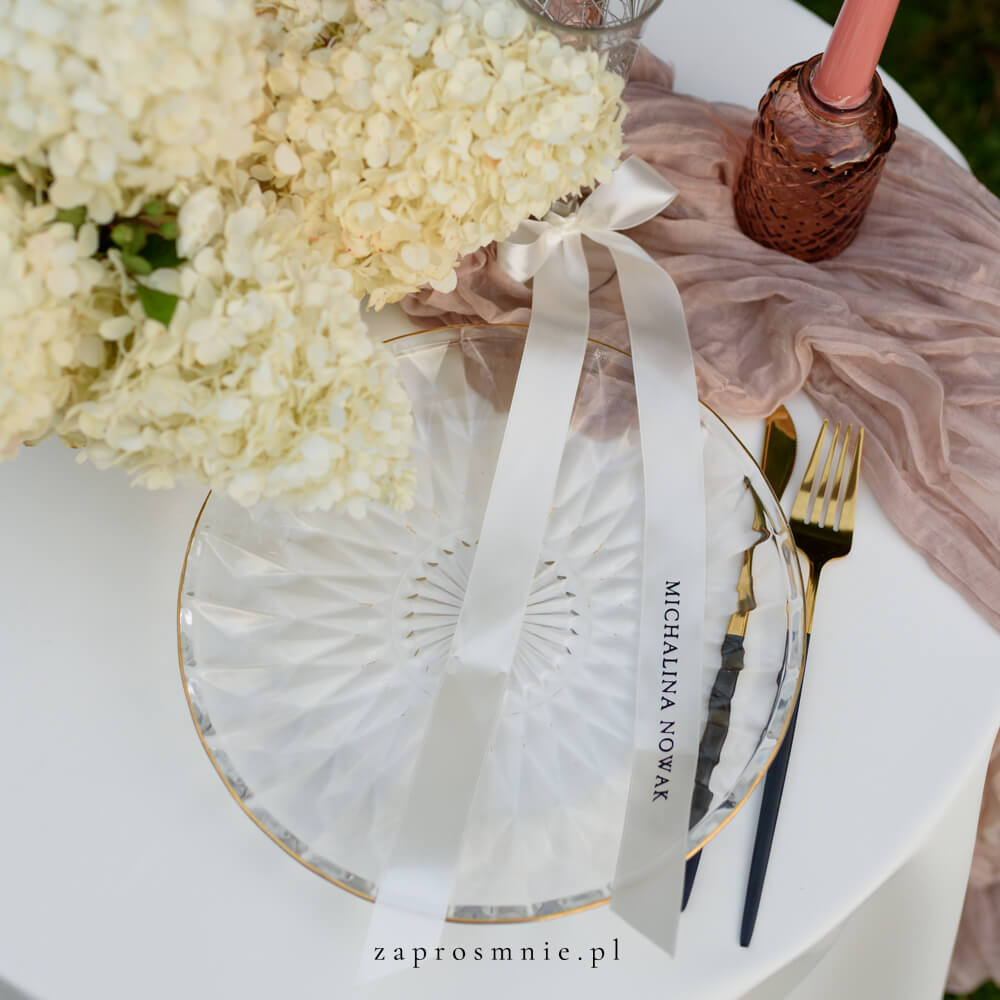 Elegancka winietka ślubna w formie śmietankowej satynowej wstążki z nadrukowanym białym, przewiązana kokardą przy kieliszku, nowoczesna i nietuzinkowa dekoracja stołu weselnego, hit Pinterest i wyjątkowa alternatywa dla klasycznych winietek.
